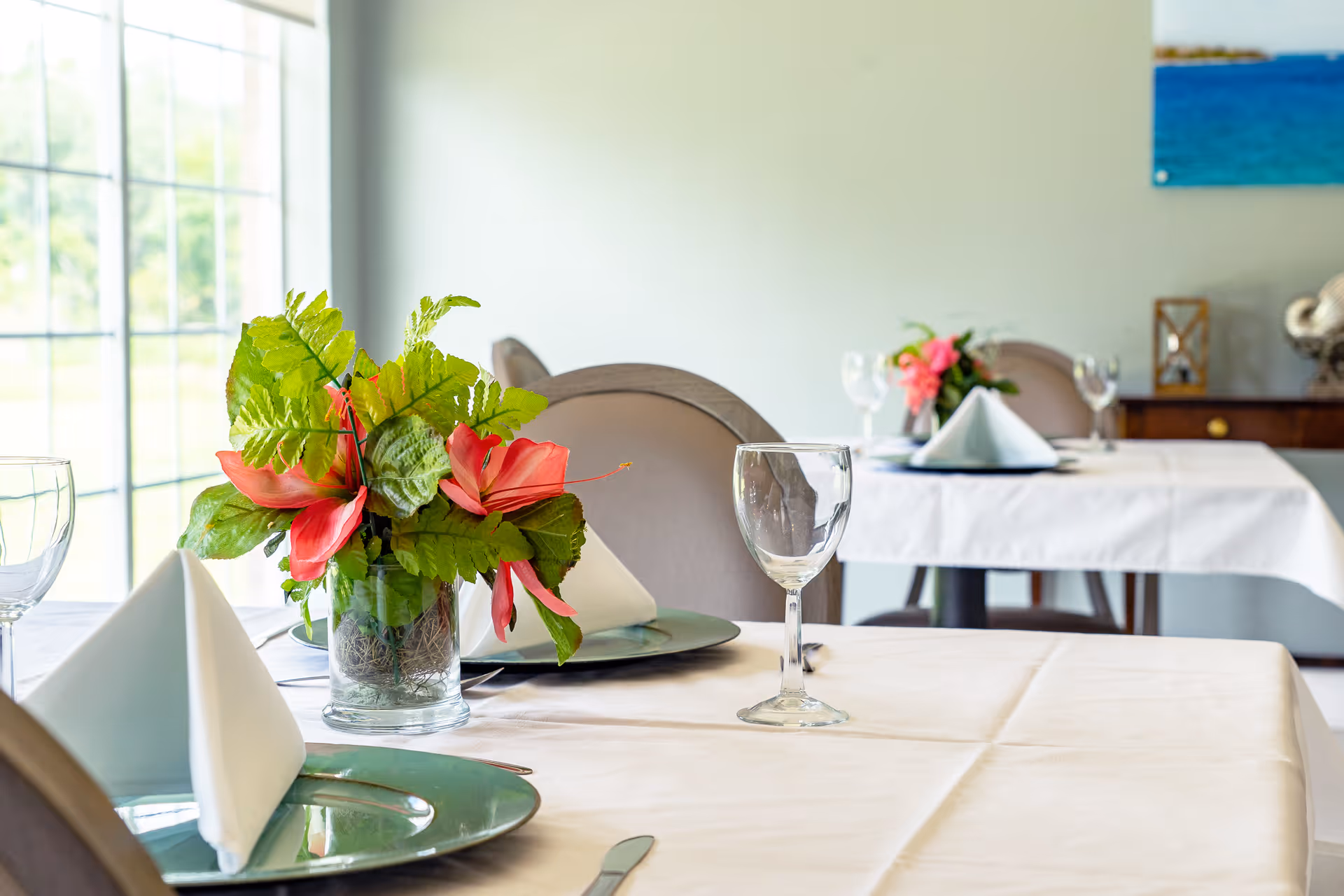 A dining room with tables set with white tablecloths, folded white napkins on plates, empty wine glasses, and floral centerpieces. The room has large windows letting in natural light and a painting on the wall.