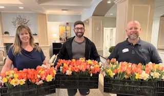 Three people standing indoors holding large black crates filled with colorful tulips and daffodils, smiling at the camera in a well-lit room with beige walls and decorative elements.