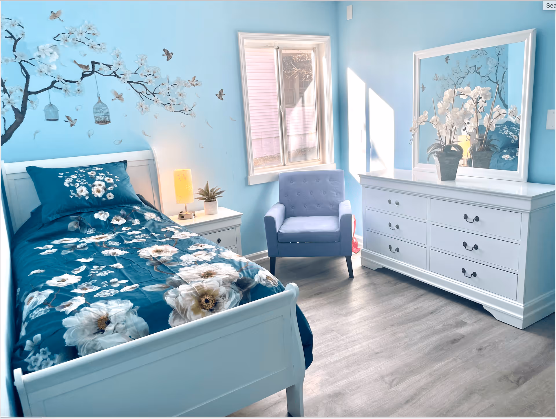 A bright bedroom with light blue walls featuring a floral and bird mural. The room contains a white single bed with a blue floral bedspread, a white nightstand with a small lamp and plant, a blue upholstered armchair, and a white dresser with a large mirror and a vase of white flowers. Sunlight streams in through a window.