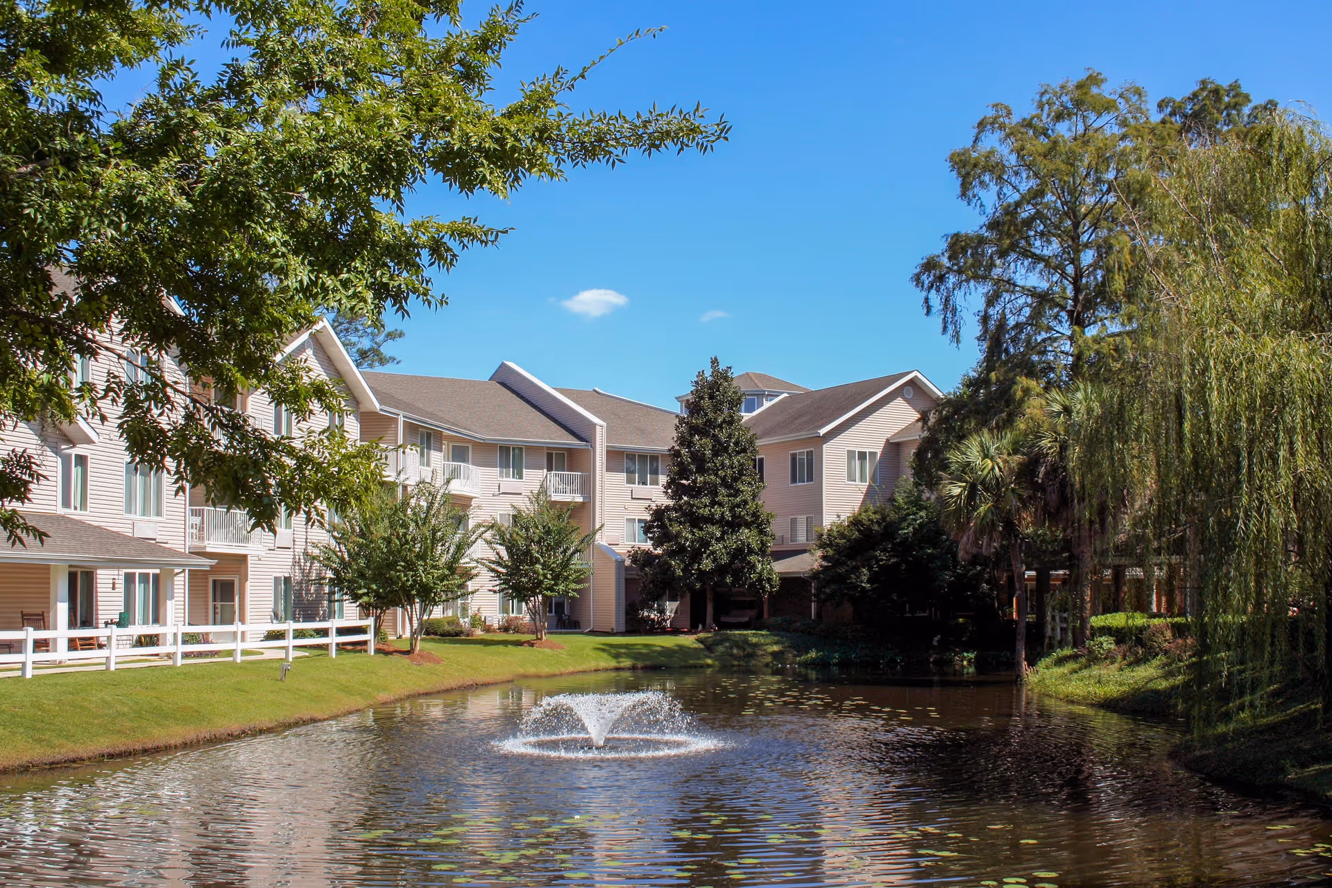 A senior living facility named River's Edge By Barclay House with beige multi-story buildings surrounding a pond with a water fountain. The area is landscaped with green grass, trees, and shrubs under a clear blue sky.