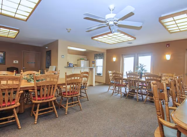 A spacious dining room with multiple wooden tables and chairs with red cushions. The room has beige walls, carpeted floors, ceiling fans, and large fluorescent light fixtures. There is a small kitchen area visible in the background with a counter and appliances, and double glass doors letting in natural light.