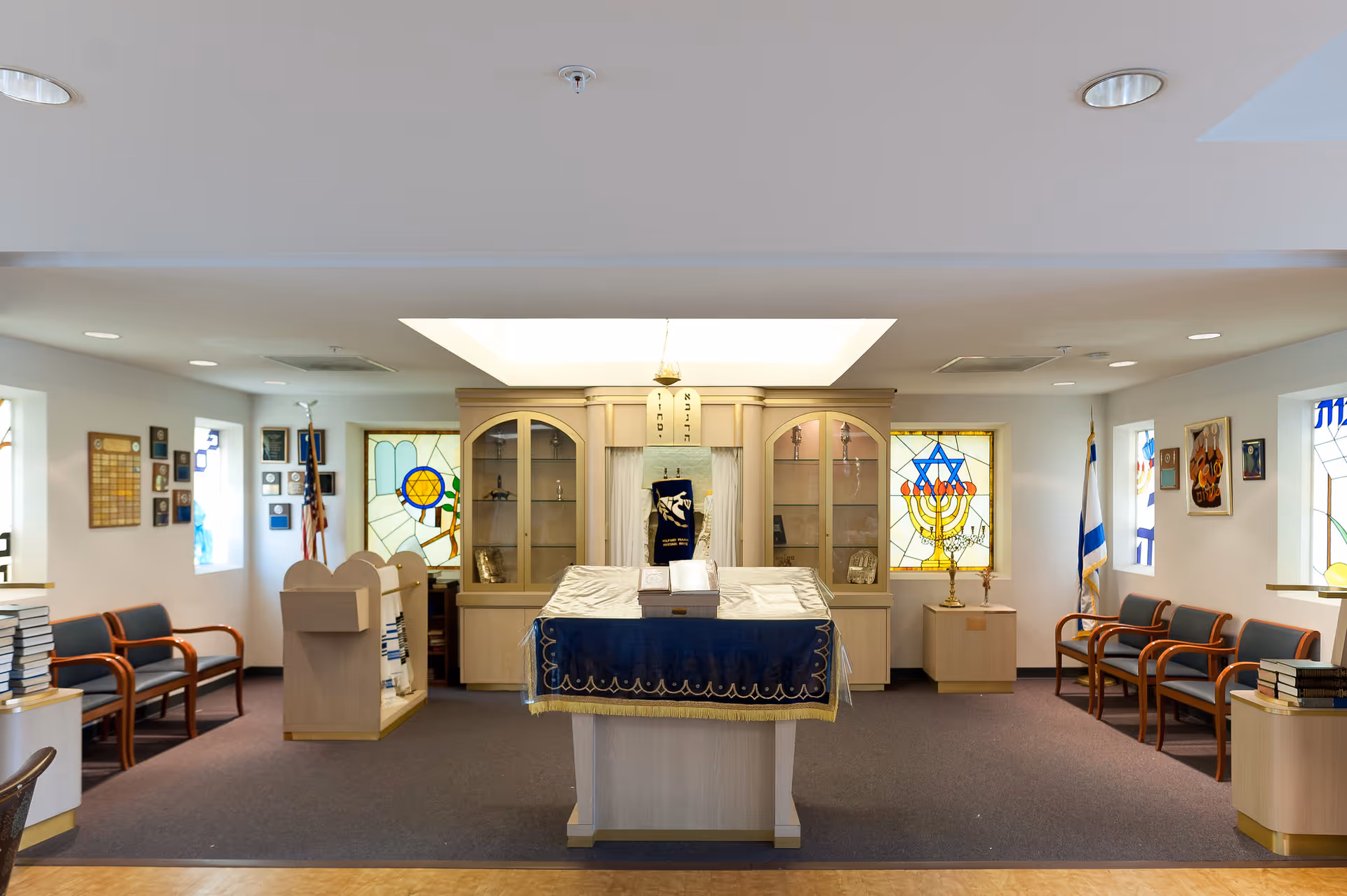 Interior view of a small synagogue or prayer room with a central reading table covered with a blue cloth, flanked by wooden cabinets with glass doors. Stained glass windows featuring Jewish symbols such as the Star of David and a menorah are visible on the walls. Chairs are arranged along the sides of the room, and various plaques and flags are displayed on the walls.