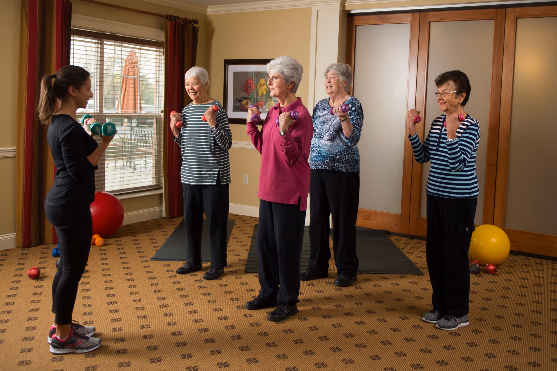 A group of four elderly women participating in a light exercise class led by a younger female instructor in a well-lit room. Each person is holding small dumbbells and smiling while exercising. The room has large windows with curtains, a carpeted floor, and exercise mats and balls in the background.