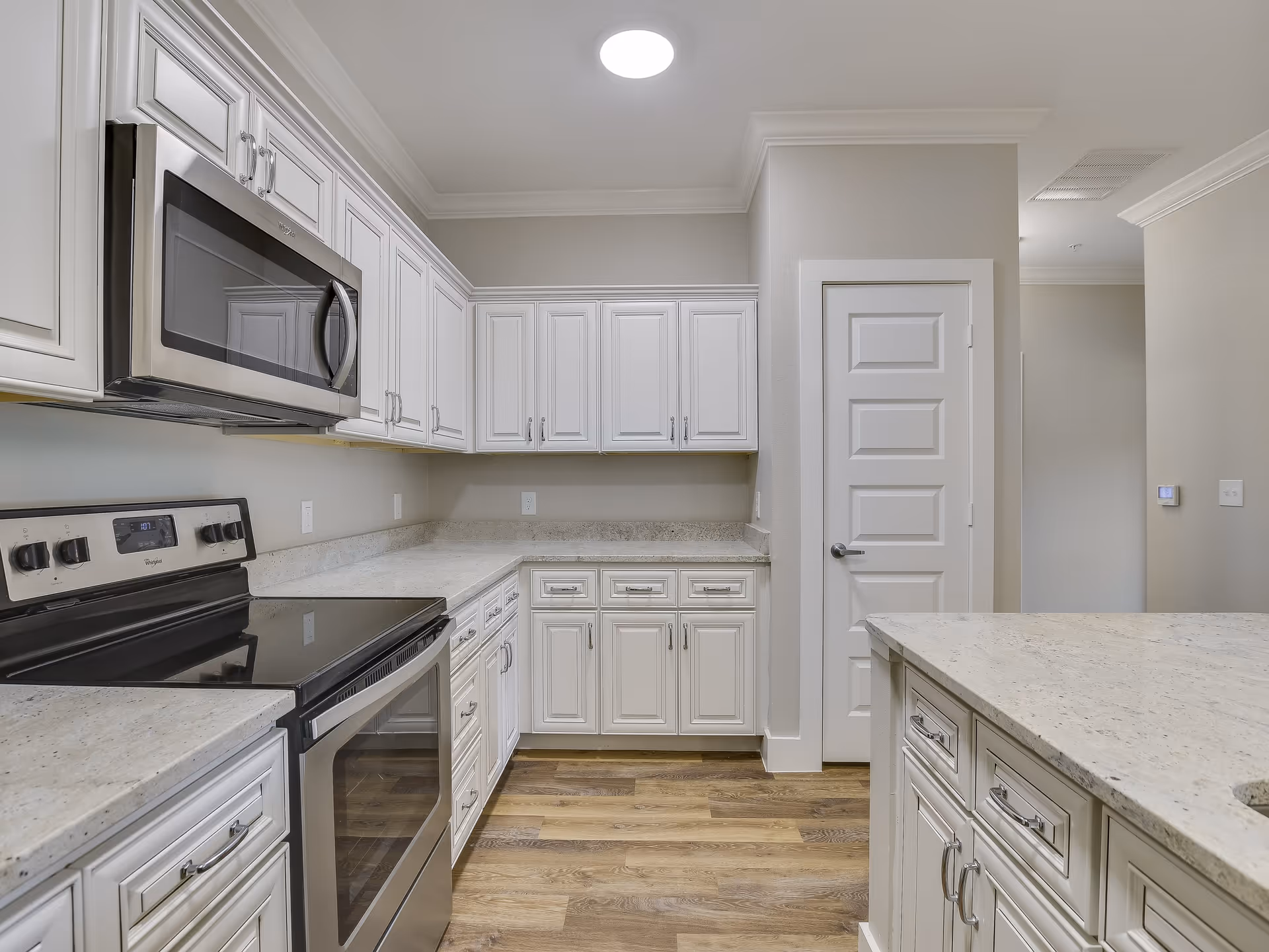 Modern kitchen with white cabinetry, stainless steel microwave and oven, light granite countertops, and wood flooring. There is a white door and a ceiling light fixture visible.