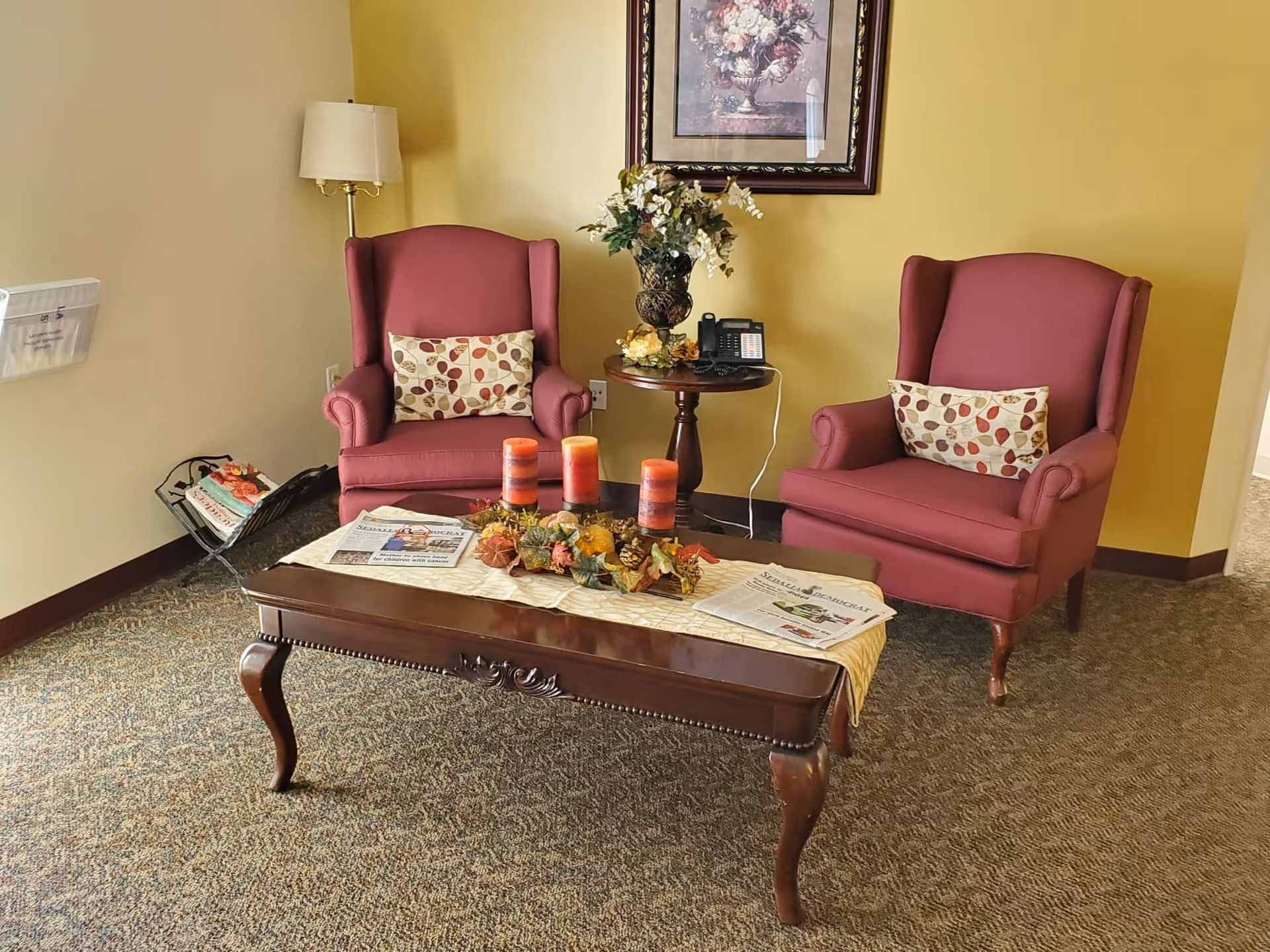 A cozy seating area with two red upholstered armchairs, each with a decorative pillow featuring a leaf pattern. Between the chairs is a small round wooden table with a floral arrangement and a telephone. In front of the chairs is a wooden coffee table with three orange candles, a fall-themed centerpiece, and two newspapers. A floor lamp stands behind the left chair, and a framed floral painting hangs on the yellow wall. The carpet is patterned in neutral tones.