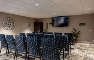 A small conference or presentation room with several rows of patterned blue chairs facing a wall-mounted flat screen TV. The room has beige walls, a patterned carpet, a decorative wall piece resembling a large gear, a small cabinet, and two potted plants.