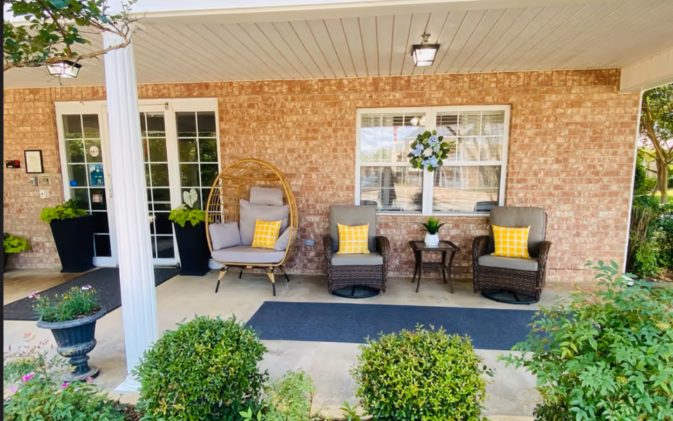 Covered outdoor patio area with brick wall background featuring two cushioned wicker chairs with yellow checkered pillows, a small table with a potted plant between them, and a cushioned hanging chair with pillows. There are potted plants on either side of a glass door entrance, a white column, and green shrubs in the foreground.