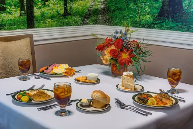 A dining table set with plates of food including a bread roll with butter, a slice of cake, a plate with vegetables and fish, a plate with sliced meats and boiled eggs, and glasses of iced tea. There is a floral centerpiece with red, orange, and yellow flowers. The background shows a wall with a nature-themed mural of green trees and foliage.