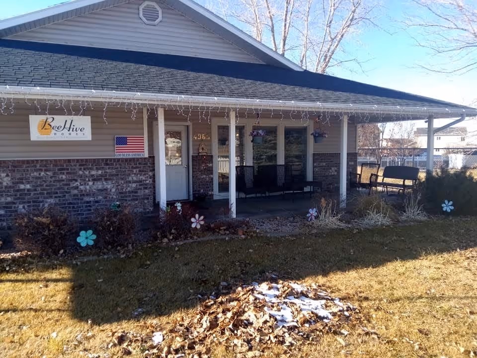 Exterior front view of Bee Hive of Roy facility showing a covered porch with chairs and hanging flower pots. The building has a brick lower half and beige siding upper half with a dark shingled roof. There is a sign with the Bee Hive logo and an American flag with the words 'God Bless America' on the wall. The lawn in front has some fallen leaves and small decorative flower stakes.