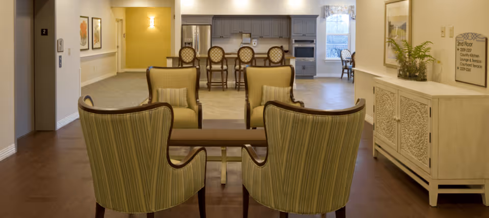 A cozy common area in a senior living facility featuring four green striped armchairs arranged around a small wooden table. In the background, there is a kitchen area with gray cabinets, a countertop with four high chairs, and a window letting in natural light. The walls are decorated with framed artwork and a sign indicating directions to the 2nd floor amenities.