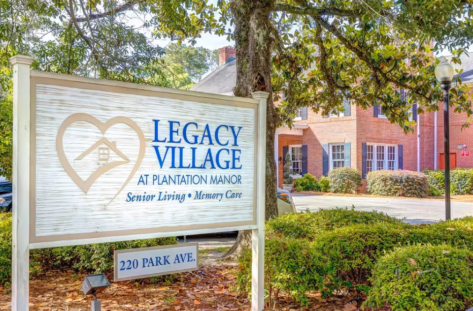 Sign for Legacy Village at Plantation Manor in front of a brick senior living building with trees and landscaped shrubs.