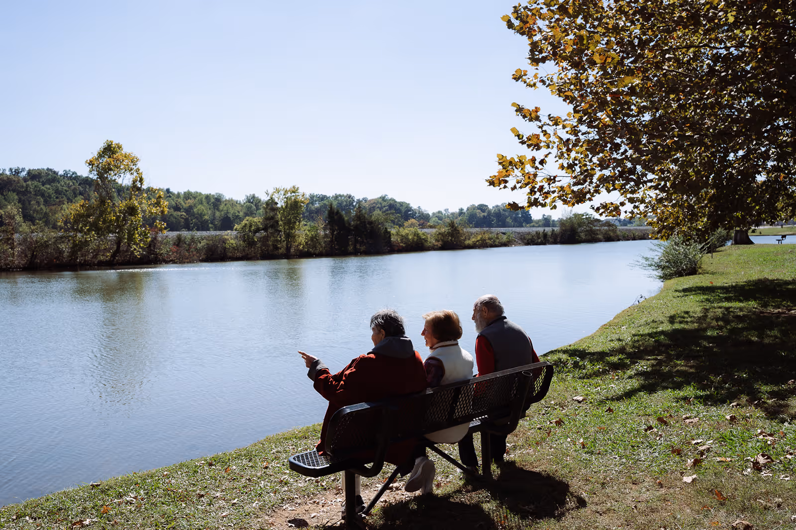 Three older adults sit on a bench by a calm lake with one person pointing toward the water under a clear sky.