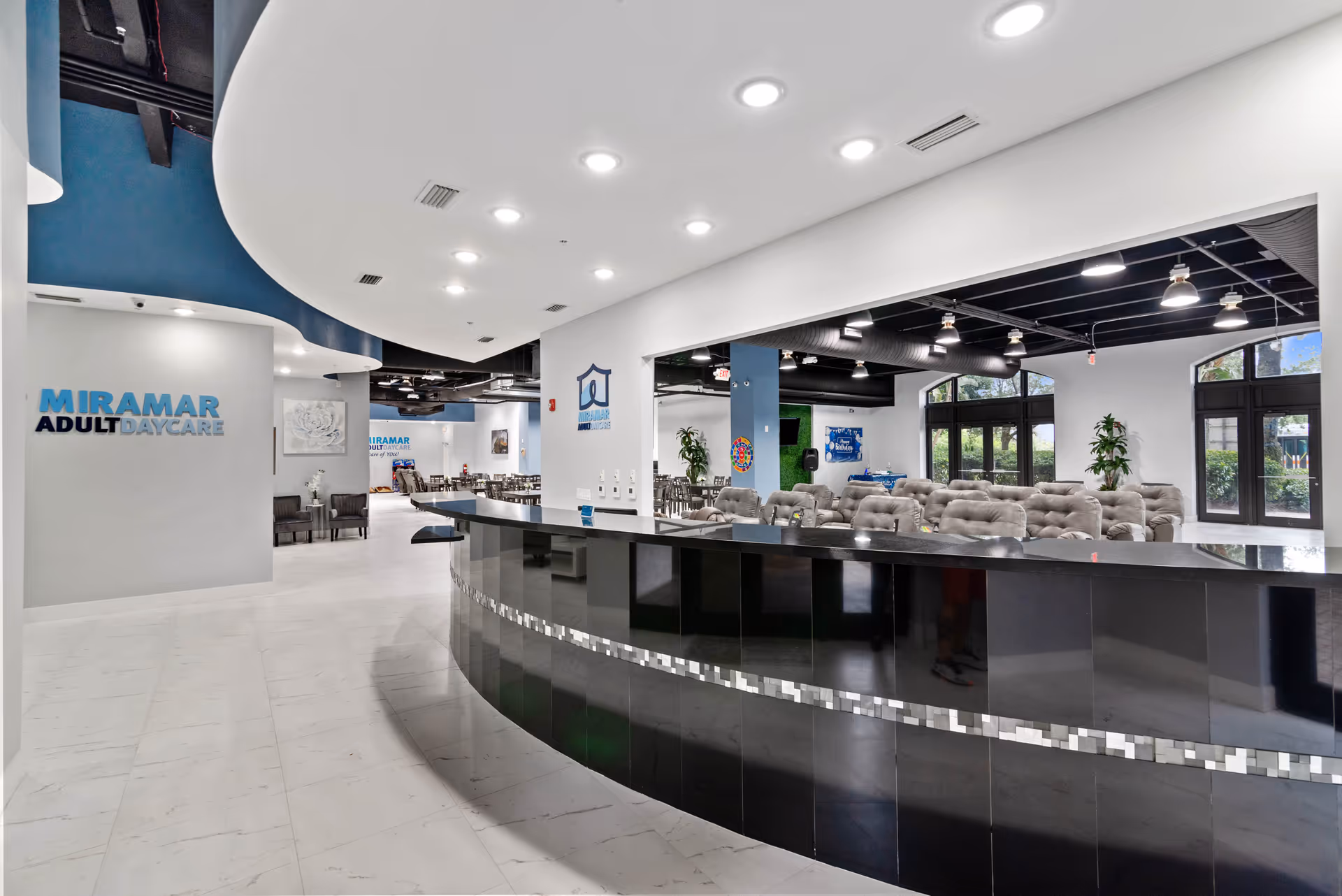 Interior view of Miramar Adult Day Care featuring a modern reception desk with a glossy black finish and a curved design. The space has white walls, a white tiled floor, and a ceiling with recessed lighting. In the background, there are multiple seating areas with cushioned chairs and tables, large windows letting in natural light, and decorative plants.