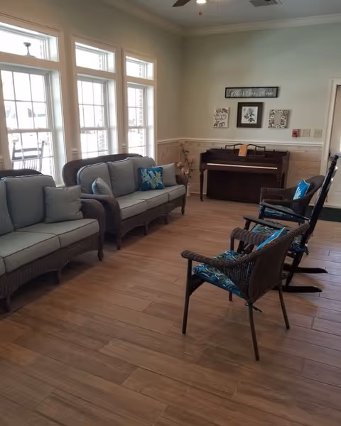A cozy living room area with wicker furniture including two sofas and two chairs with blue cushions. There is a piano against the wall with framed artwork above it. Large windows let in natural light, and the floor is wooden.