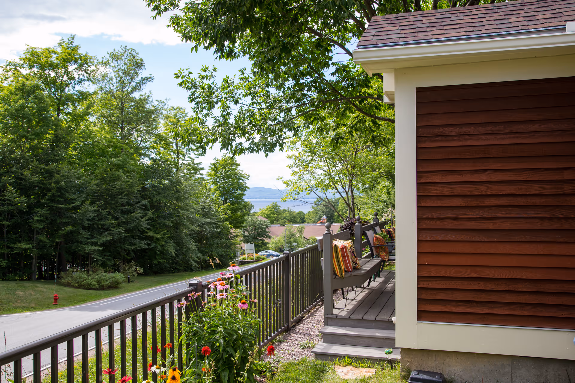 View of a porch with benches and colorful cushions overlooking a road, surrounded by green trees and flowering plants, with a glimpse of distant hills under a partly cloudy sky.