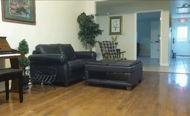 A seating area with a black leather loveseat, matching ottoman, a rocking chair and a piano on hardwood floors leading to a hallway.