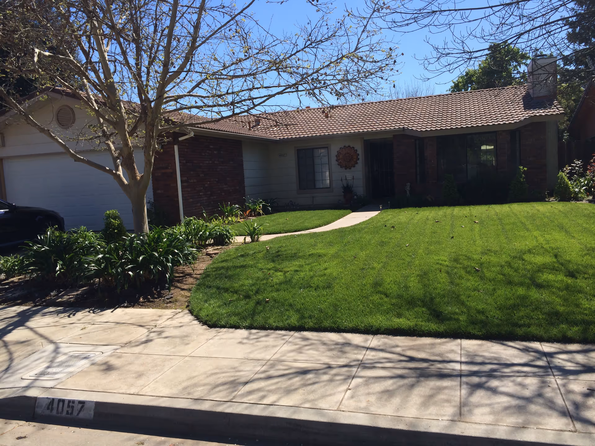 Single-story suburban house front with a tiled roof, brick facade, green lawn, and a sidewalk.