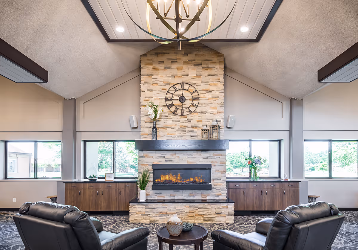 A cozy senior living common area featuring a stone fireplace with a modern electric fire, a large round clock mounted above it, and decorative vases and lanterns on the mantel. Two black leather armchairs face the fireplace with a small round table between them. Large windows on either side of the fireplace let in natural light, and wooden cabinets run along the walls beneath the windows. The ceiling is vaulted with a large circular chandelier hanging down.