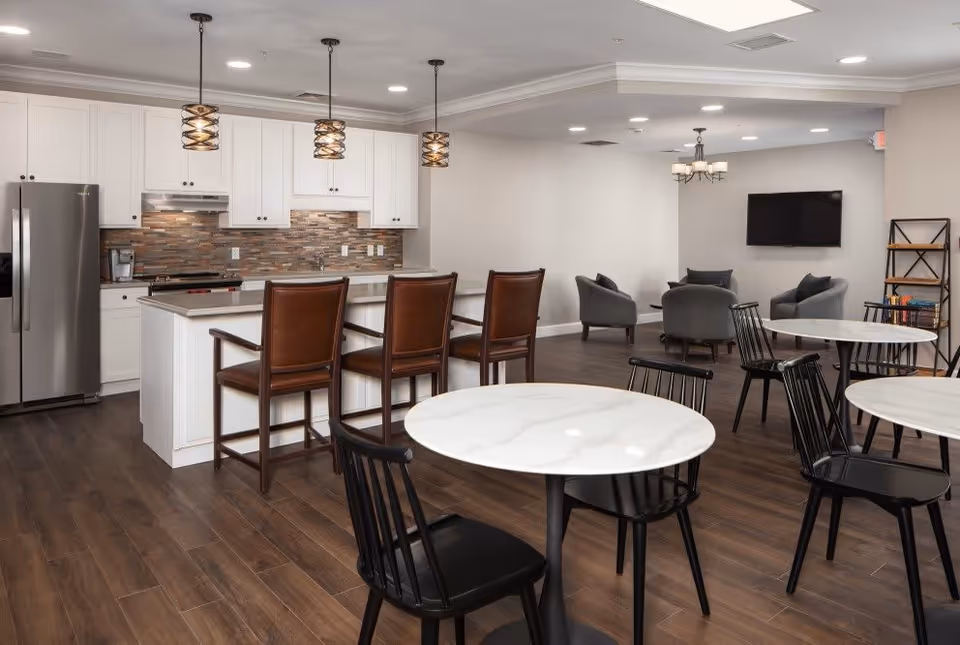 A modern communal space featuring a kitchen area with white cabinets, a stainless steel refrigerator, and a kitchen island with three brown leather bar stools. The room also includes several round tables with black chairs, a seating area with gray armchairs, a wall-mounted TV, and a bookshelf. The flooring is dark wood, and the lighting includes pendant lights over the island and a chandelier in the seating area.