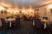 A warmly lit dining room with multiple round tables set for a meal. Each table is covered with a white tablecloth and decorated with floral centerpieces and tableware. The room features blue upholstered chairs, patterned carpet, wall sconces with lights, and framed artwork on the walls.