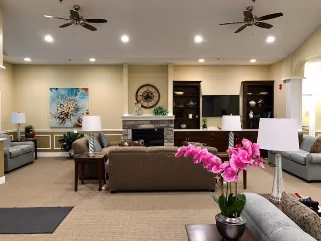 A spacious living room with beige walls and carpeted floor, featuring a central stone fireplace with a clock above it. The room is furnished with multiple sofas and armchairs arranged around the fireplace, side tables with lamps, a large flat-screen TV on a wooden cabinet, and decorative shelves. A vibrant pink orchid in a pot is placed on a table in the foreground.