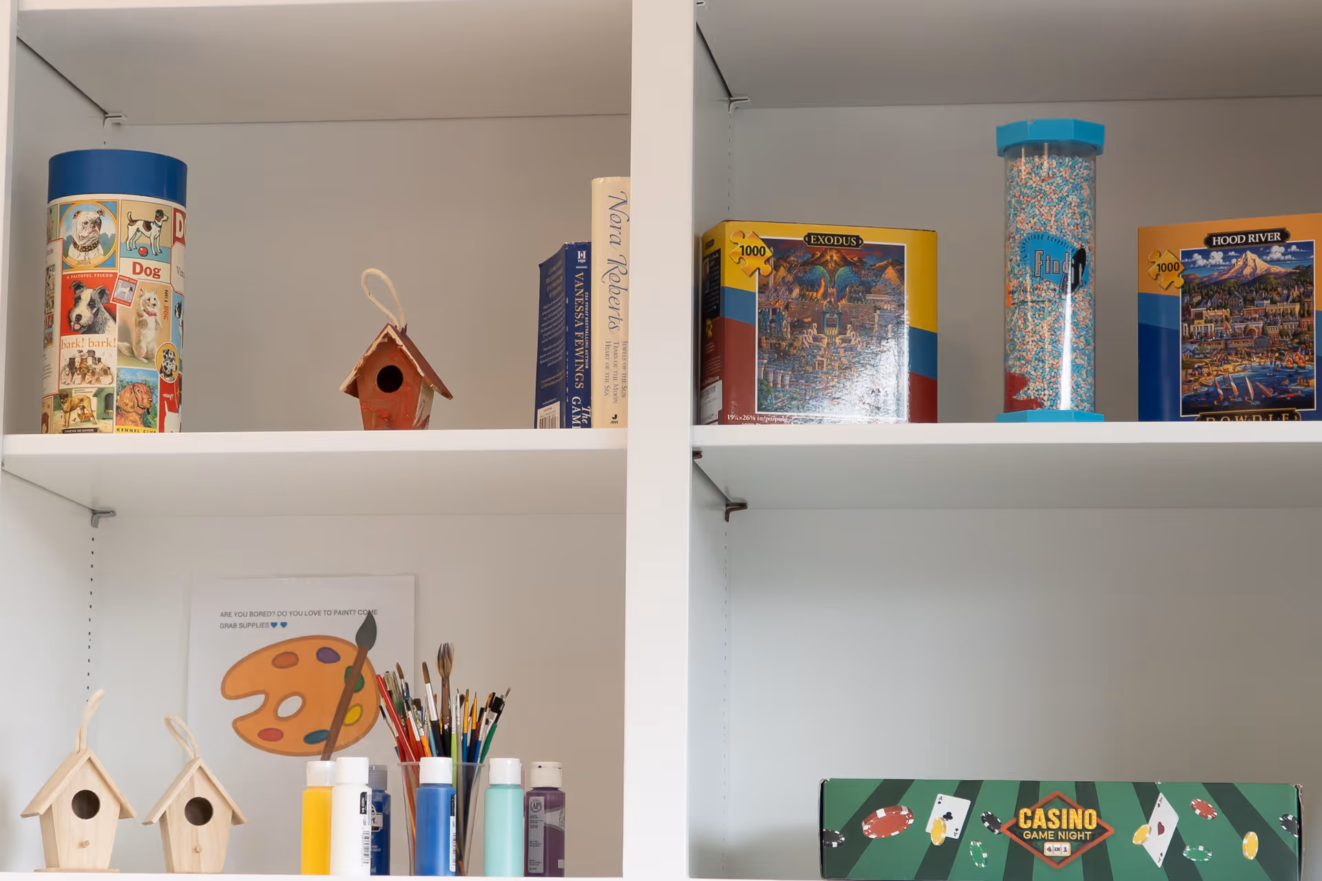 White shelves holding various items including a cylindrical container with dog images, small wooden birdhouses, paint bottles and brushes, books, puzzle boxes, a container filled with colorful beads, and a board game box labeled 'Casino Game Night'.