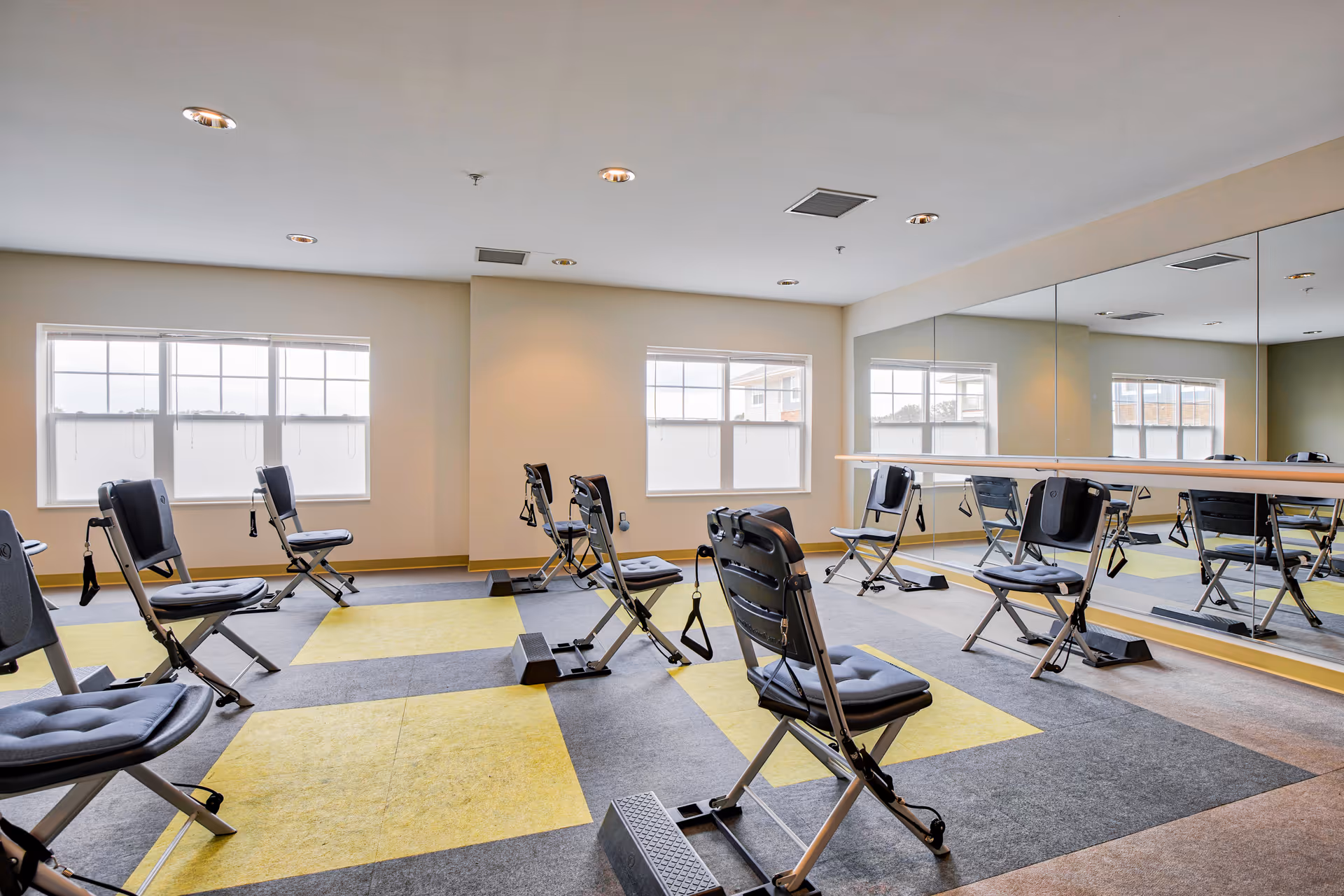 A bright exercise room with multiple padded folding chairs equipped with resistance bands, large windows letting in natural light, and a wall covered with mirrors reflecting the room.