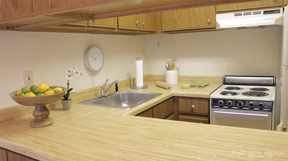 A kitchen area with wooden cabinets and light-colored countertops. The kitchen features a stainless steel sink with a faucet, a white electric stove with four burners, a paper towel holder, a small potted orchid, a wooden bowl filled with lemons and limes, and some kitchen utensils in a holder. A wall clock is mounted above the sink.