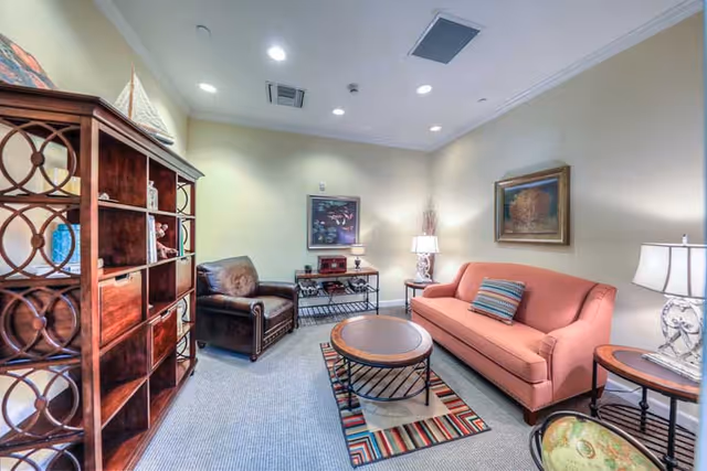 A cozy living room with a coral-colored sofa, a brown leather armchair, a round coffee table on a colorful striped rug, two side tables with lamps, a wooden shelving unit, and framed artwork on the walls.