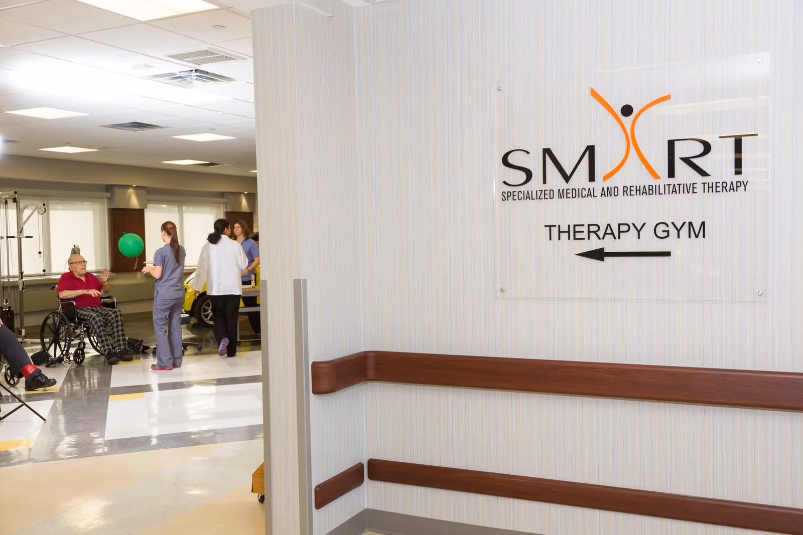 Hallway entrance to a therapy gym with a wall sign reading 'SMART Therapy Gym' and staff helping a patient in a wheelchair visible inside.