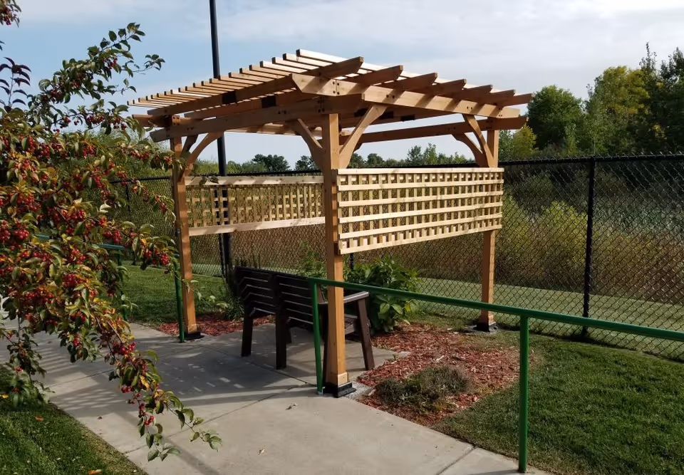 A wooden pergola with lattice sides situated on a concrete path in a garden area. Underneath the pergola is a dark bench. The area is surrounded by green grass, a black chain-link fence, and trees in the background. There is a green handrail along the path and a bush with red berries on the left side.