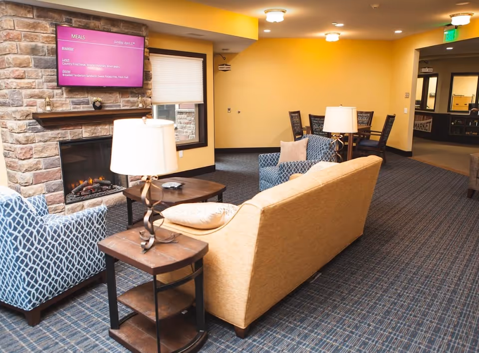 A cozy living room area in a retirement community with a stone fireplace, a mounted TV displaying meal options, a beige sofa, two blue patterned armchairs, wooden side tables with lamps, and a dining table with chairs in the background. The walls are painted yellow and the floor is carpeted.