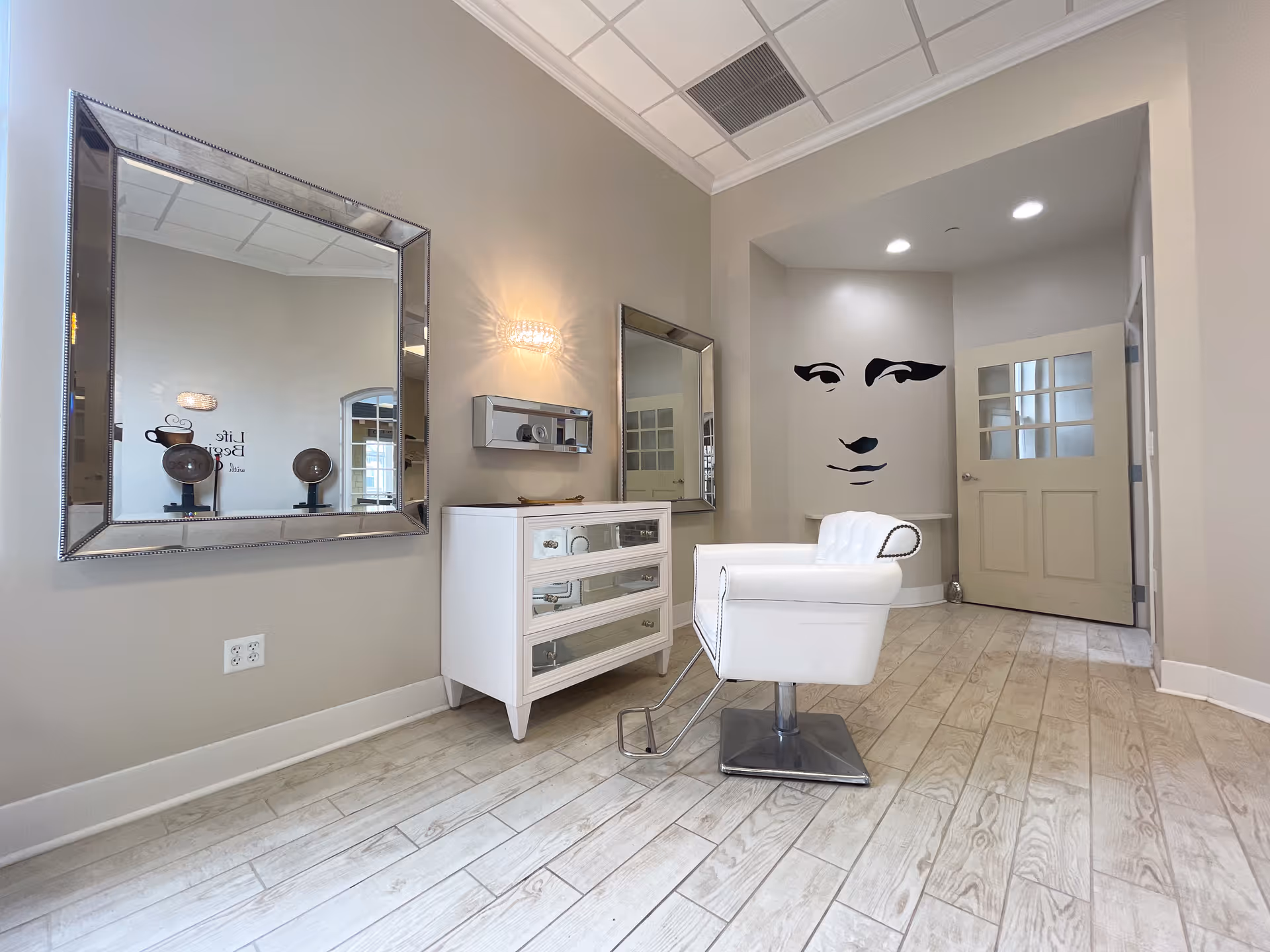 A modern salon area with a white salon chair in front of a white dresser with mirrored drawers. Two large square mirrors hang on the light gray walls, and a wall light fixture is mounted between them. In the background, there is a wall with a stylized black and white face artwork and a door with glass panels. The floor is light-colored wood.