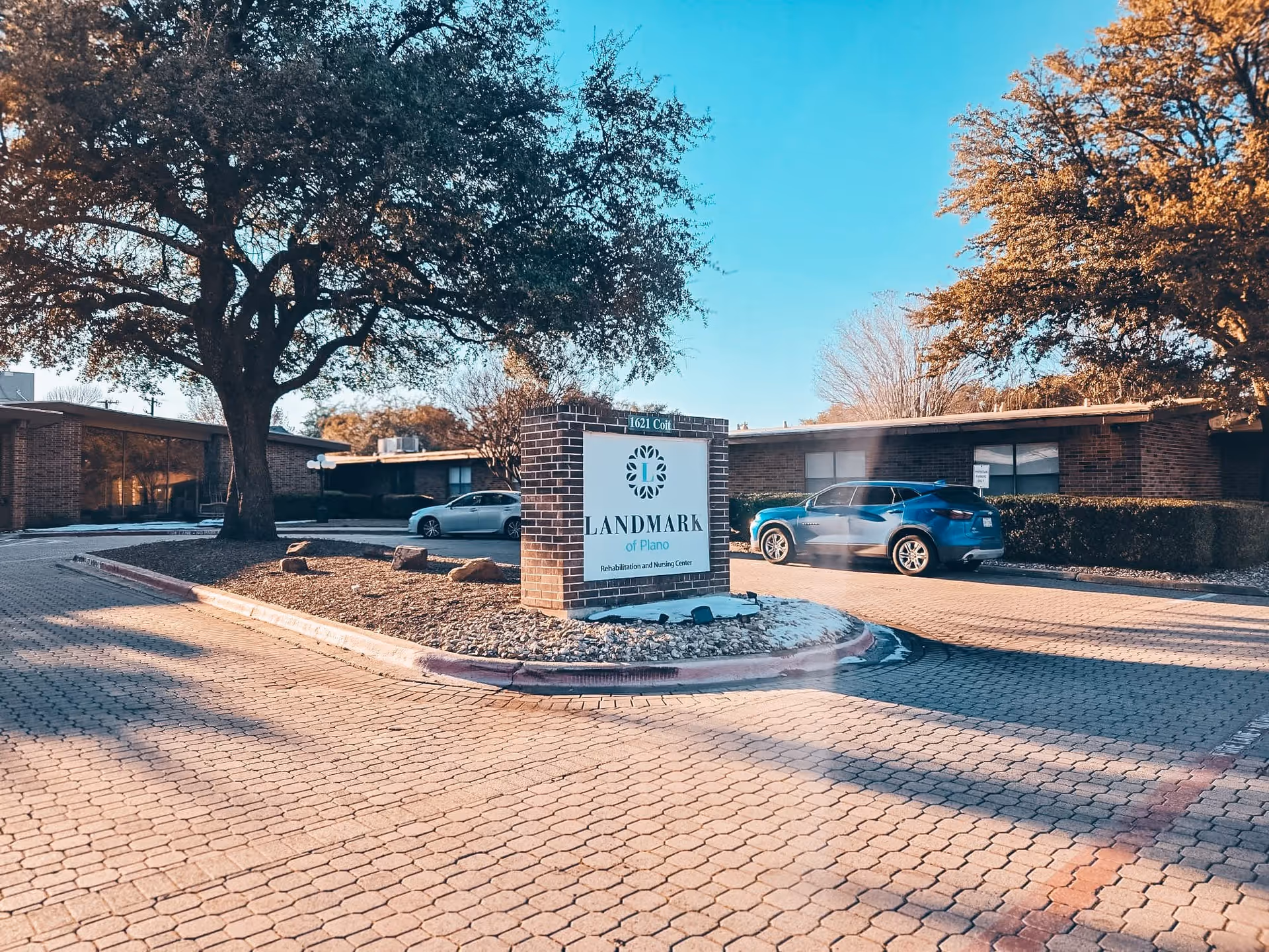 Exterior view of the Landmark of Plano Rehabilitation and Nursing Center showing a brick sign with the facility name, a paved driveway, parked cars, and trees under a clear blue sky.