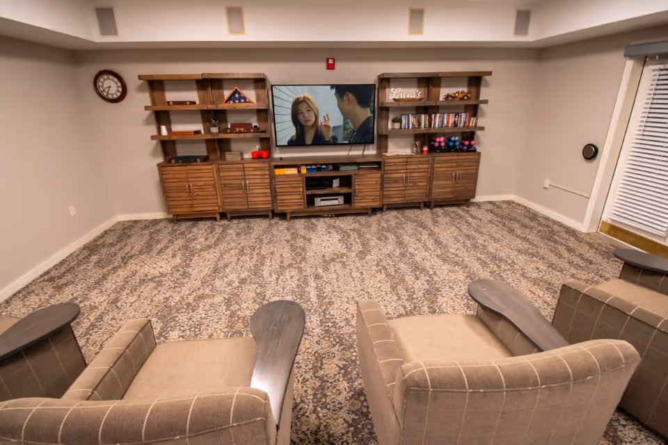 A cozy living room with two beige armchairs facing a wooden entertainment center with shelves and cabinets. A flat-screen TV is mounted on the wall above the center, displaying a scene from a show or movie. The room has patterned carpet flooring, a wall clock on the left, and a window with blinds on the right.