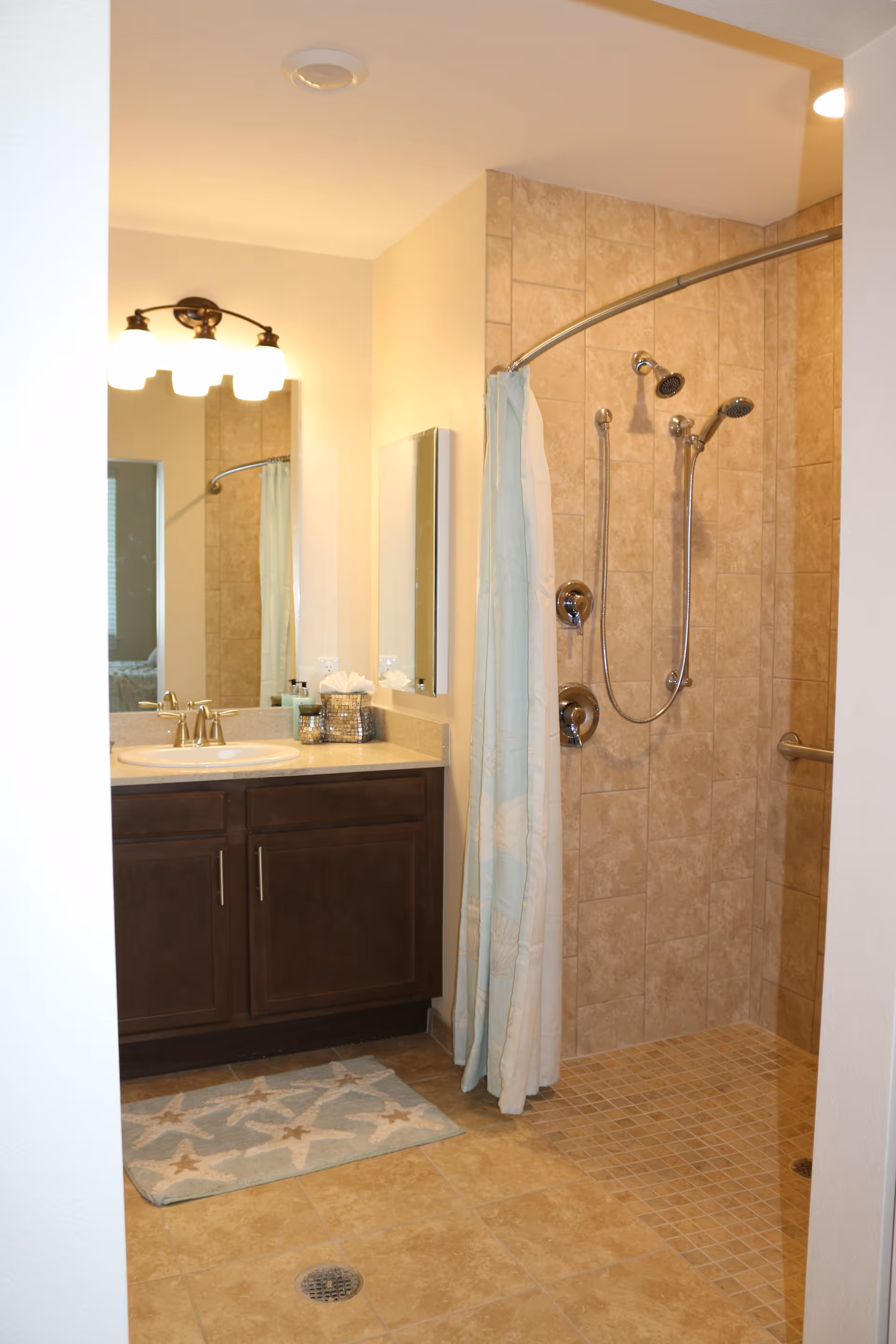 A bathroom featuring a tiled walk-in shower with two showerheads and a curved shower curtain rod with a light blue and white curtain. Next to the shower is a vanity with a sink, countertop, and dark wood cabinets below. Above the vanity is a mirror and a light fixture with three bulbs. A starfish-patterned bath mat is placed on the tiled floor in front of the vanity.