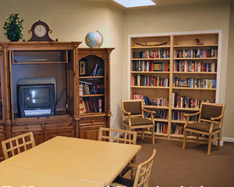 A cozy room with wooden furniture including a table with chairs, a wooden entertainment center holding a small TV and a clock, a globe on top, and bookshelves filled with books. Two wooden chairs are placed near the bookshelves against beige walls.