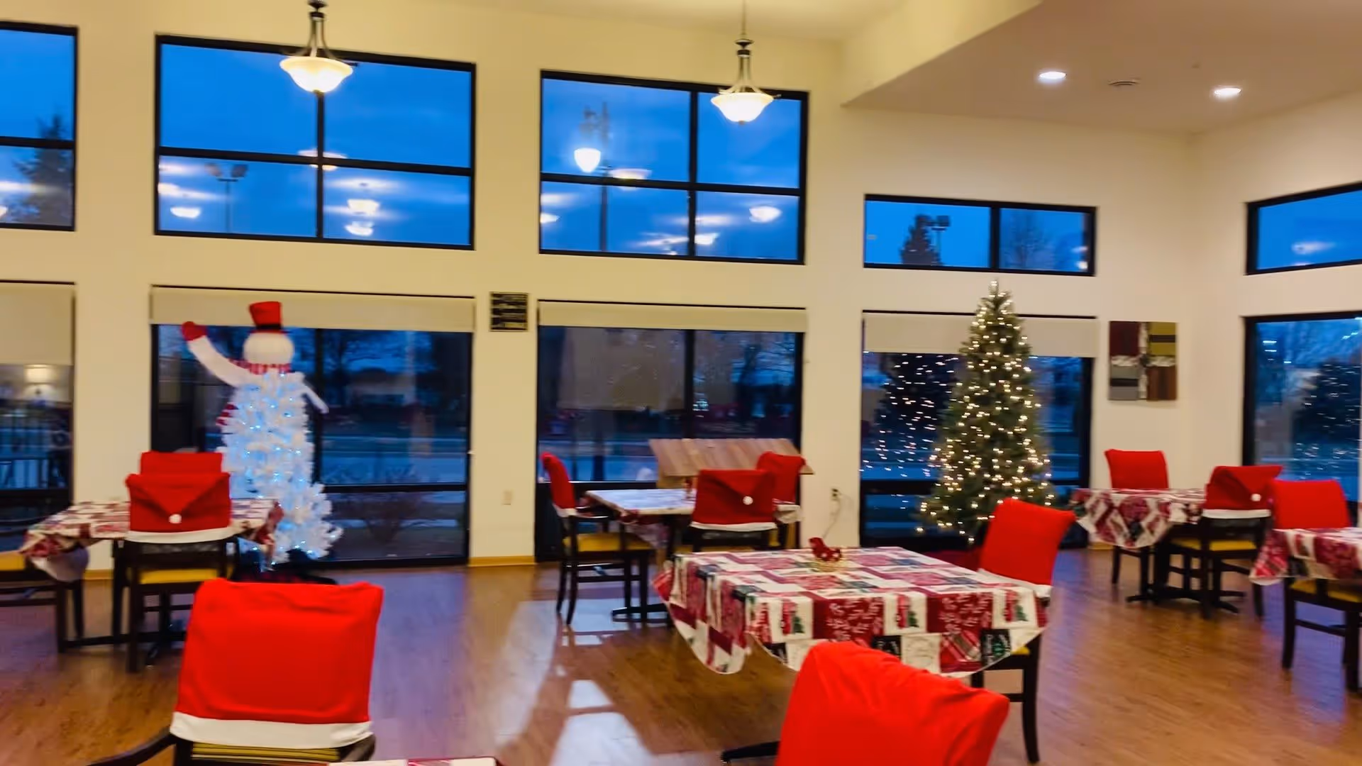 Interior of a senior living facility dining room decorated for Christmas with tables covered in festive tablecloths and chairs draped in red covers. A lit Christmas tree and a white snowman decoration with a red hat are visible near large windows showing an evening sky outside.