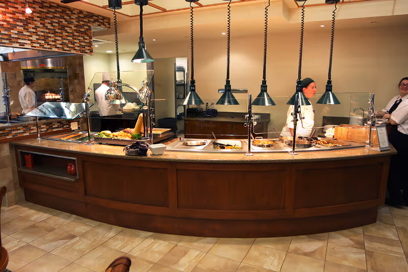 Curved buffet serving counter with food trays and staff under hanging heat lamps in a dining room.