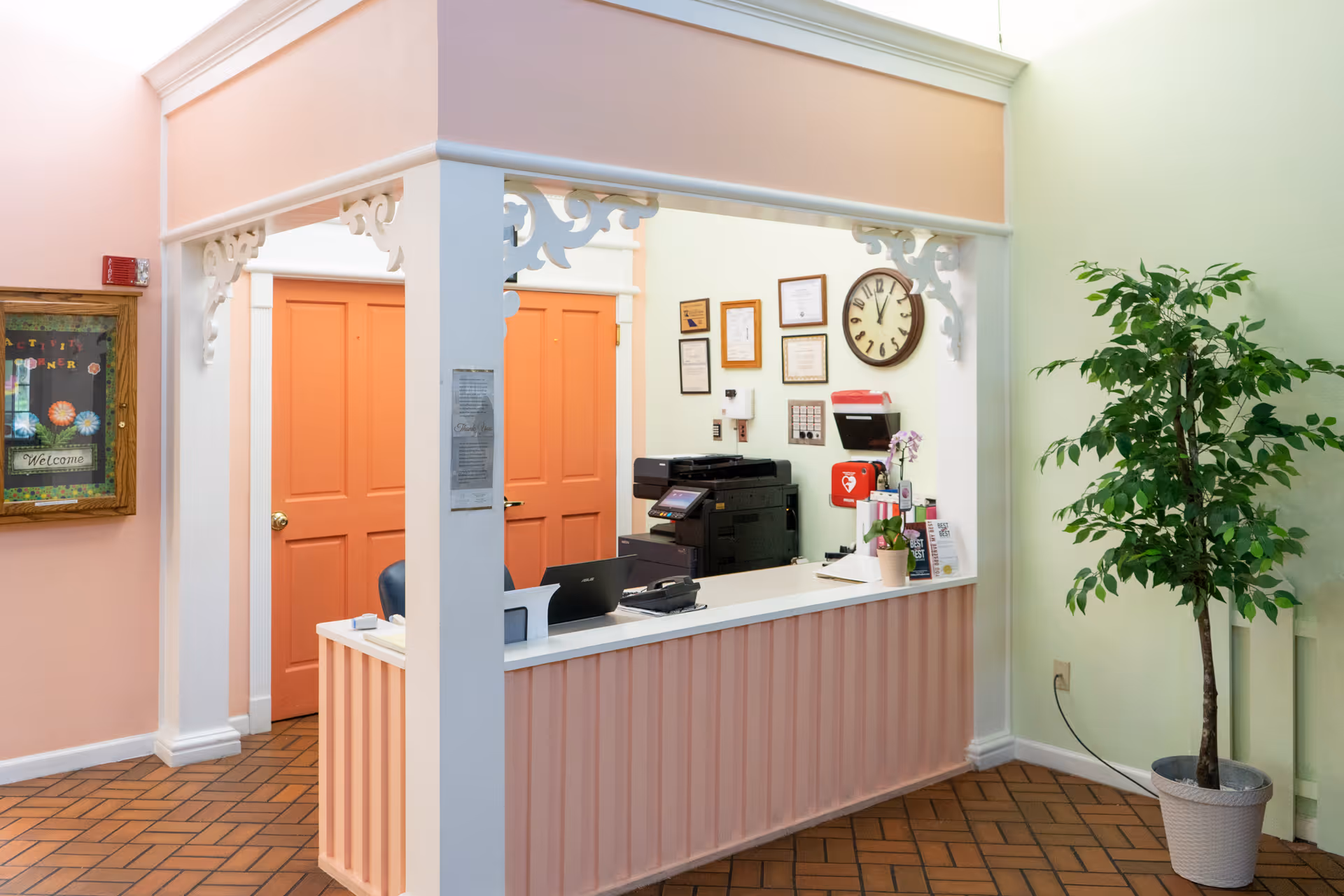 Reception area with a peach-colored front desk, decorative white trim, and two orange doors in the background. A clock and framed certificates hang on the wall behind the desk. A potted plant is placed to the right of the desk, and a bulletin board with a welcome message is on the left wall.