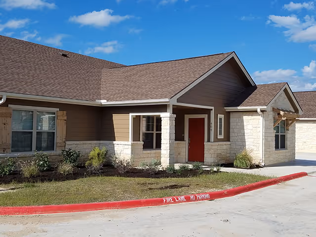 Exterior view of a single-story building with brown siding and stone accents, featuring a red front door, windows with wooden shutters, and a landscaped area with small plants. The sky is blue with scattered clouds.