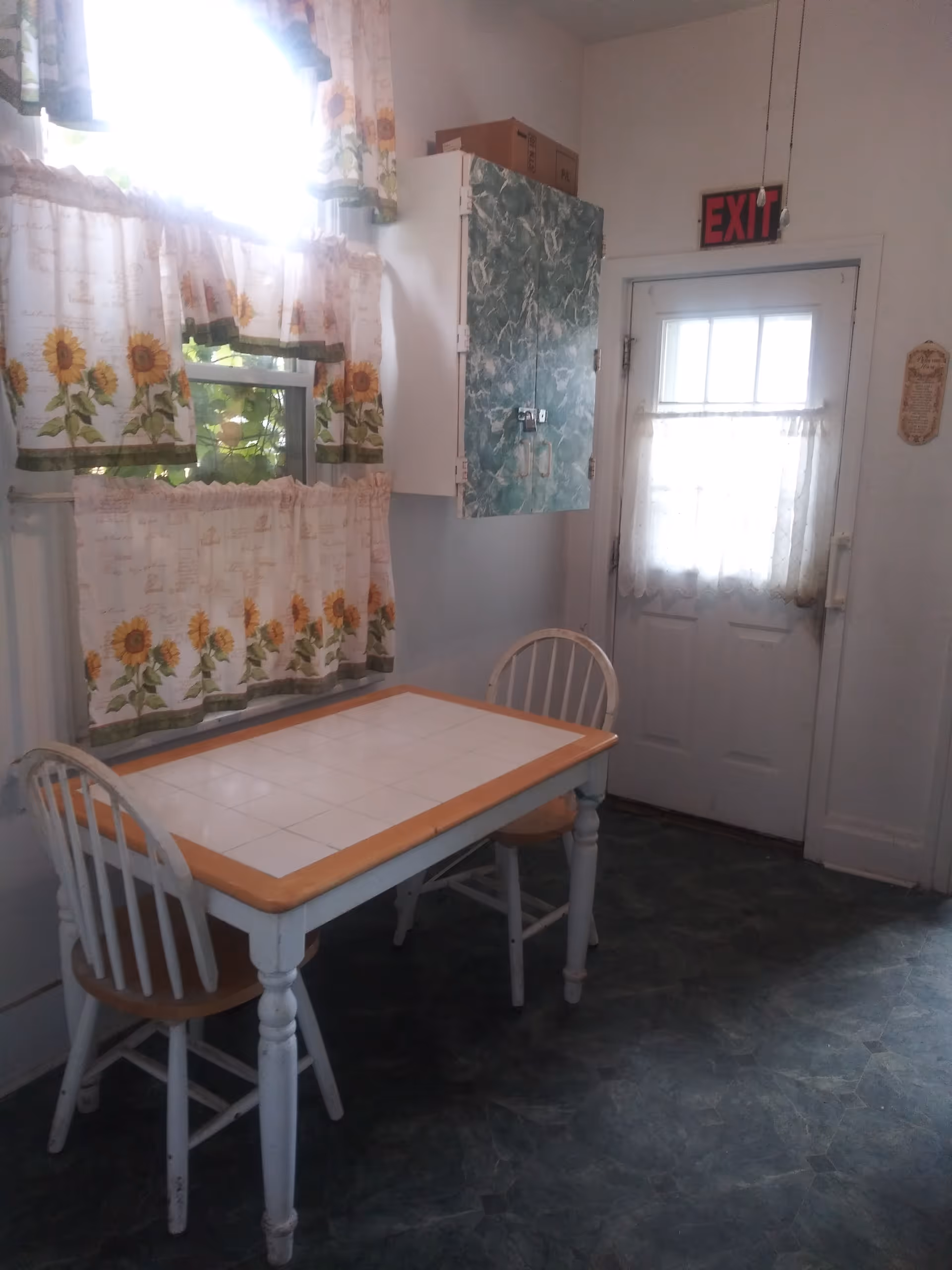 A small kitchen dining area with a white table featuring a tiled top and wooden edges, accompanied by two white wooden chairs with natural wood seats. The room has a window with sunflower-patterned curtains and a door with a lace curtain. A green marbled cabinet is mounted on the wall above the table, and an illuminated red EXIT sign is above the door.