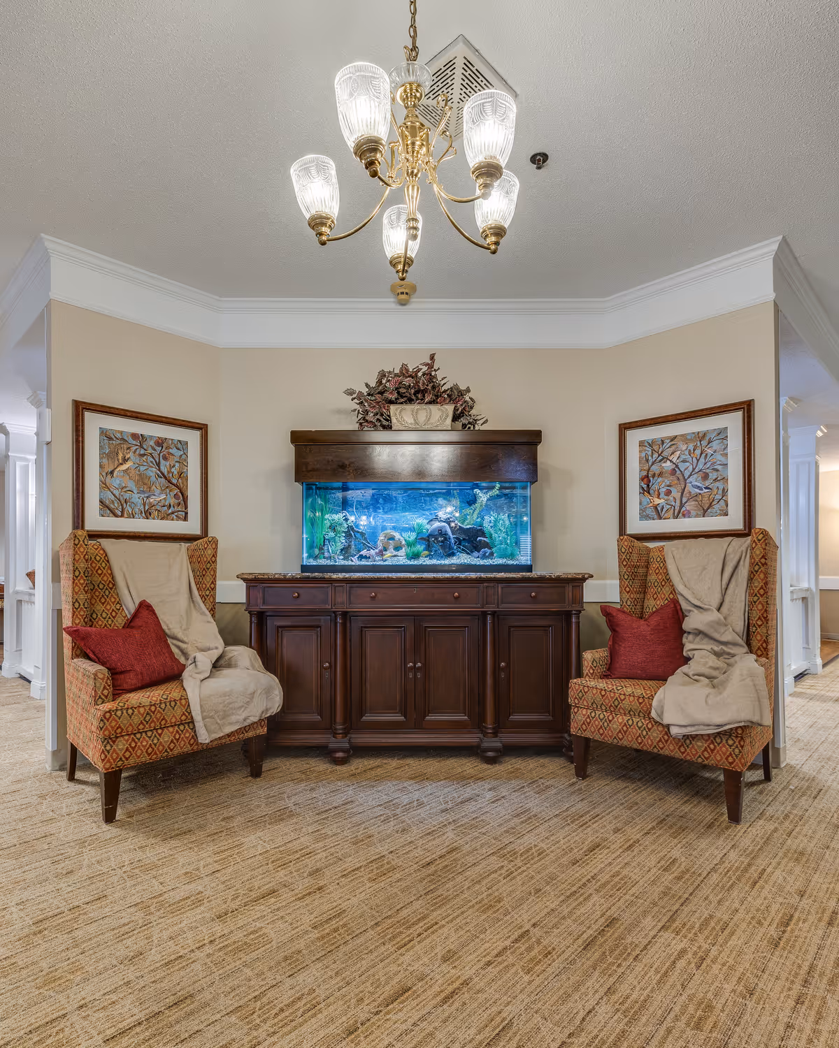 A cozy seating area in a senior living facility featuring two patterned armchairs with red pillows and beige throws, positioned on either side of a wooden cabinet with a large aquarium on top. Above the aquarium is a decorative plant, and two framed artworks hang on the beige walls behind the chairs. A brass chandelier with five glass shades hangs from the ceiling.