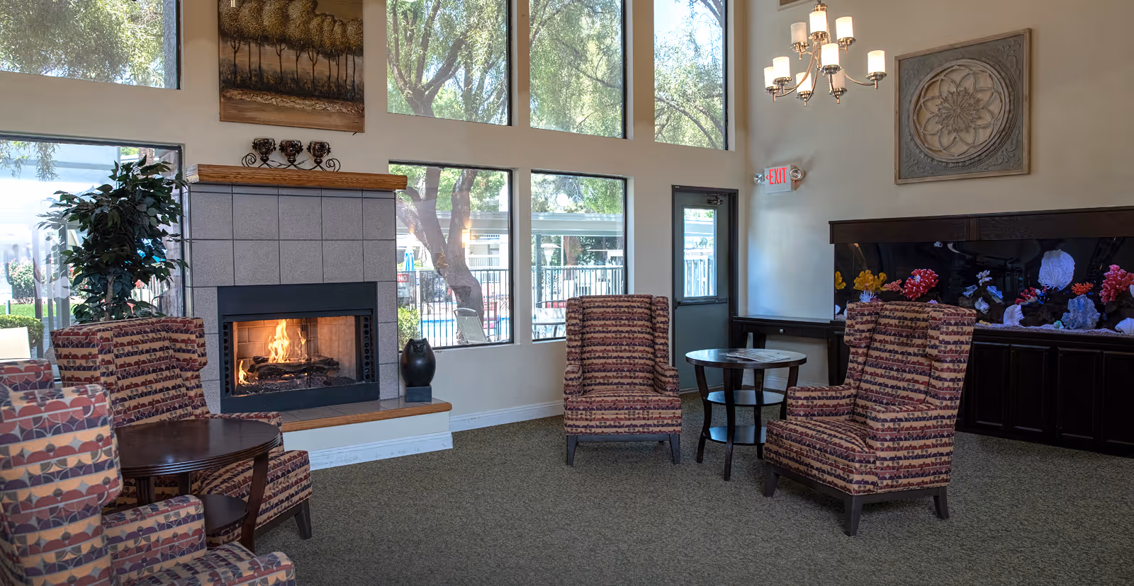 Sunlit lounge with a lit fireplace, patterned armchairs, round tables, large windows and an aquarium in a senior living facility.
