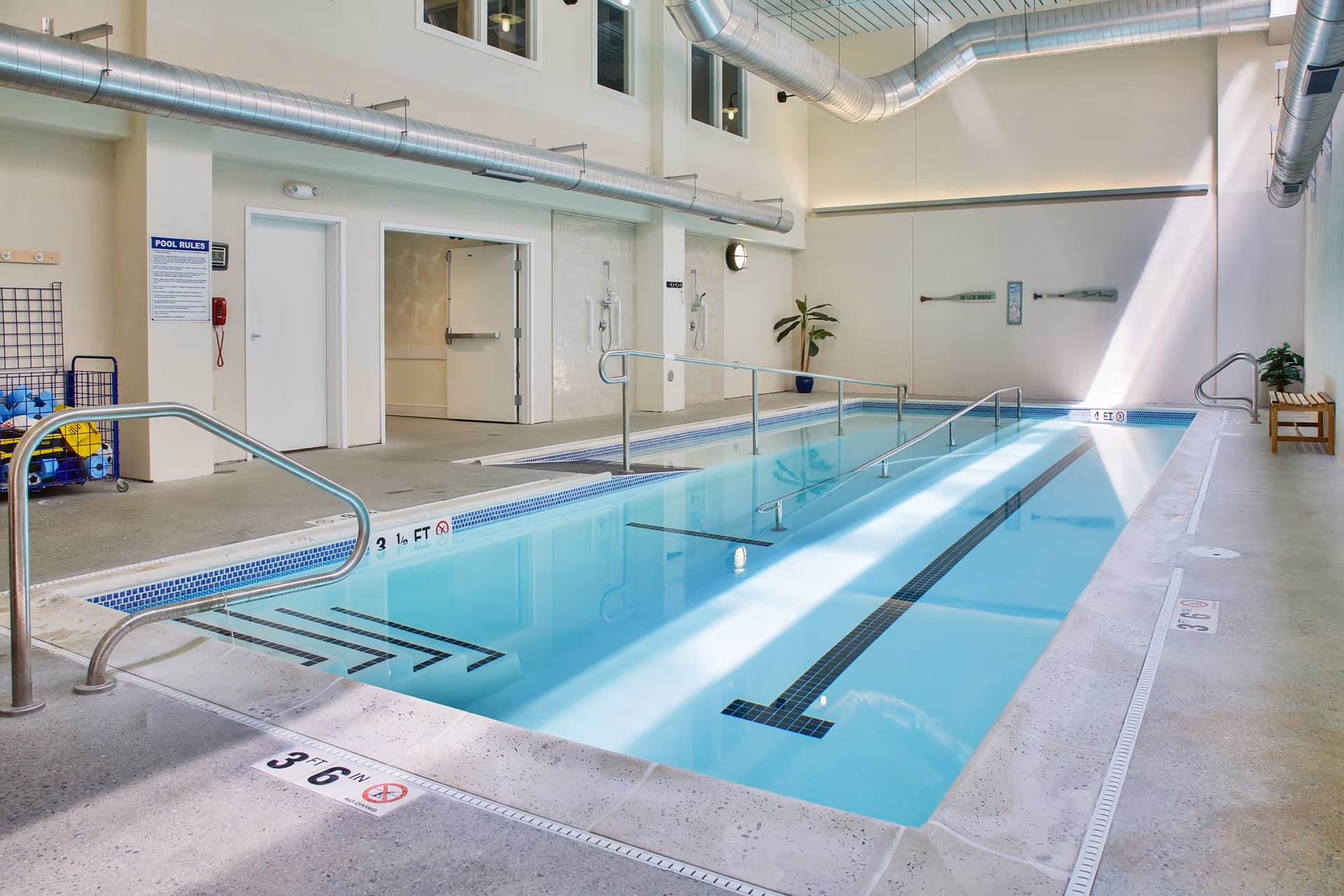Bright indoor swimming pool with handrails, lane markings, and seating along the side in a senior living facility.