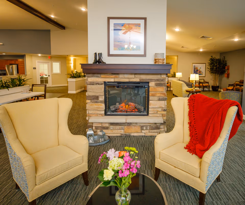 A cozy living room area in a senior living facility featuring two beige armchairs with patterned sides, one draped with a red blanket, positioned around a small round glass table with a vase of colorful flowers. A stone fireplace with a glowing fire is centered in the background, topped with decorative items and a framed picture above. The room has warm lighting and additional seating areas visible in the background.