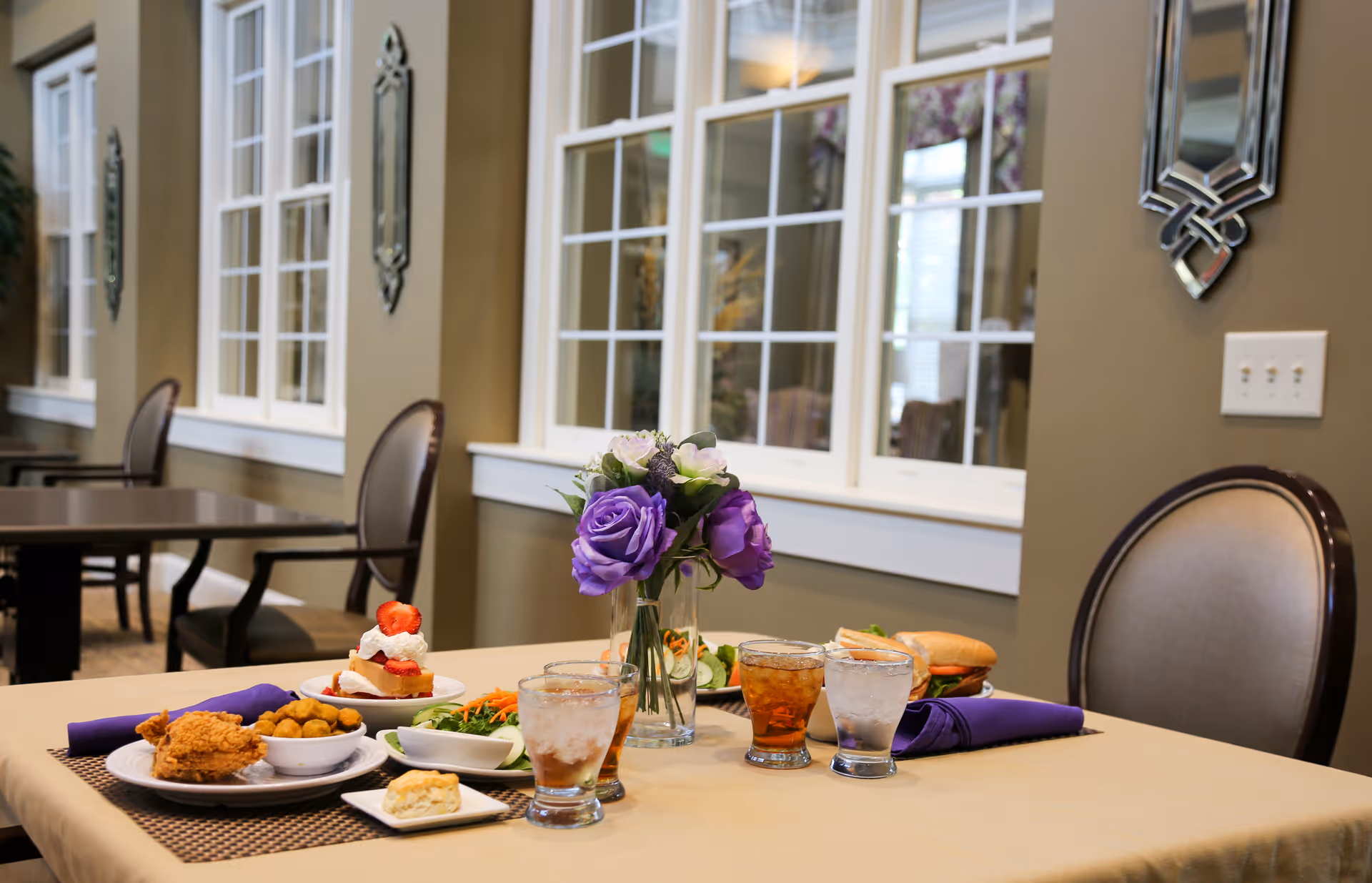 A dining table set with plates of food including fried chicken, salad, a biscuit, a sandwich, and a dessert topped with strawberries and cream. There are also three glasses with beverages and a vase with purple and white flowers. The background shows a room with large windows and decorative wall mirrors.