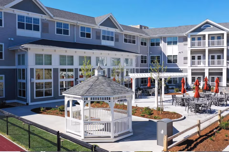 Outdoor courtyard area of a retirement community featuring a white gazebo, several tables with red umbrellas, young trees, and a multi-story building with large windows in the background under a clear blue sky.