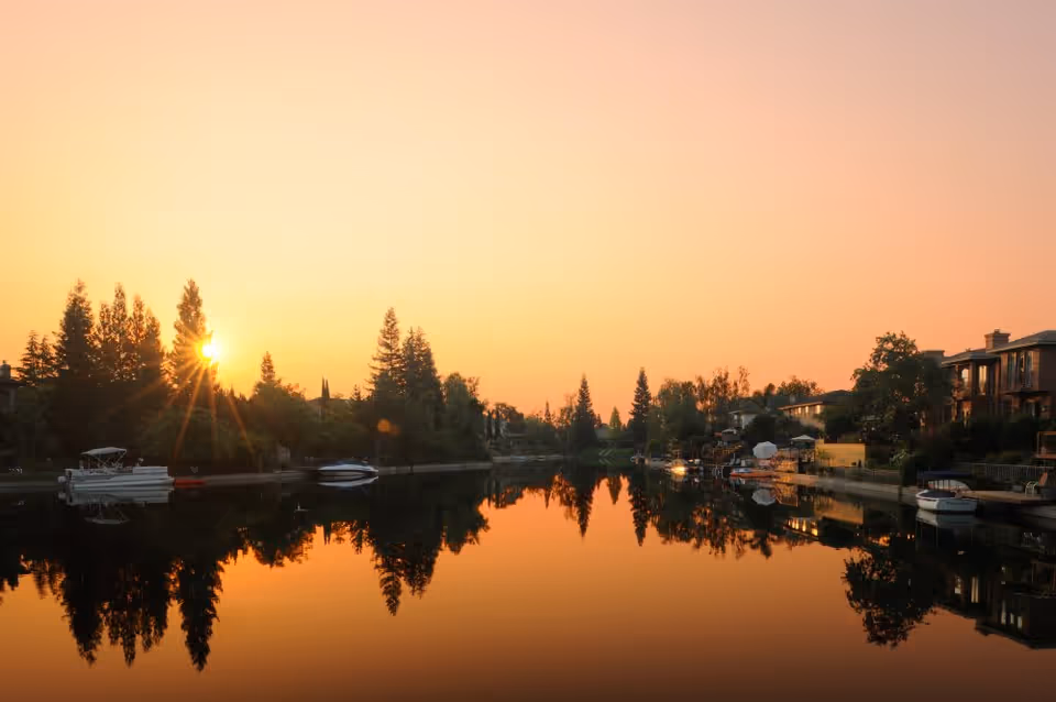Sunset over a calm river with trees, boats, and houses reflected in the water.