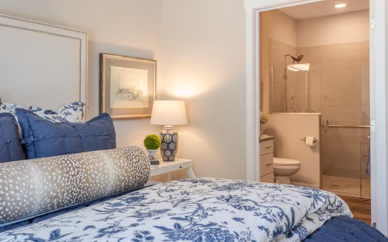 A cozy bedroom with a bed featuring blue and white floral bedding and multiple pillows. A bedside table with a decorative lamp and a small green plant is next to the bed. In the background, an open door reveals a bathroom with a toilet, a glass-enclosed shower, and beige tiled walls.